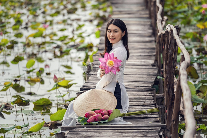 在大莲花湖维特南青山或东方的木桥上挂着粉色莲花的传统维特南帽子坐在木桥上的美丽女肖像绿色坐着敖图片