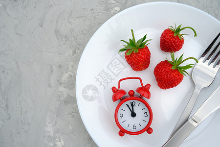 新鲜白盘上的红熟草莓浆果灰色石头背景桌上的餐具和红色闹钟特写顶视图平躺复制空间概念饮食和排毒时间或夏季菜单白盘上的红熟草莓浆果餐图片