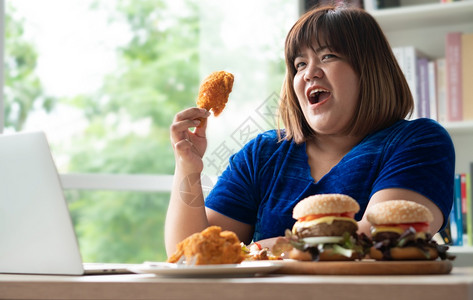 肥胖手午餐饥饿的超重女持有炸鸡木板上的汉堡包和桌披萨在家工作期间体重增加问题百食不厌症BED概念图片