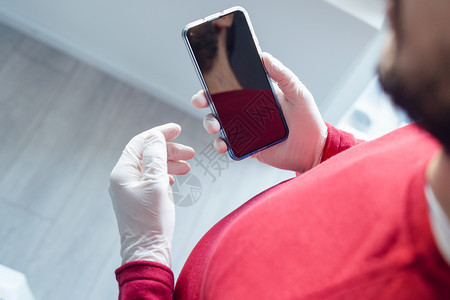疾病发短信面具成人男子在使用移动电话进行流感染期间在办公室或家中检疫时身着保护橡胶手套而站立时持有移动智能手机的成年男子顶高角度图片