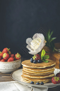 一种烤瓷炉小陶罐的糖浆以及黑木桌和色底的鲜花上面有白莓煎饼蓝和薄荷素食主义者烹饪图片