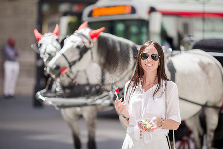 维也纳有街头食物的年轻女青在马车背景中有漂亮的马匹美丽女孩与在户外水果的美貌女孩旅游客在维也纳度假和看着在马车上的漂亮匹具历史缰图片