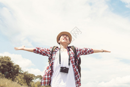 乐趣松弛身戴帽子检查衬衫相机和背包的亚洲英俊男子在夏季旅行时穿戴帽子照相机和背包他感到快乐和清新背景为蓝天和白云色的图片
