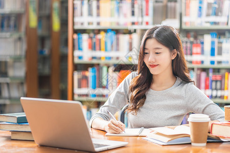 做功课和在大学图书馆或使用技术笔记本电脑的亚洲青年学生或具有各种书籍和文具架背景的各种群落返回学校从事家庭作业和使用技术笔记本电图片