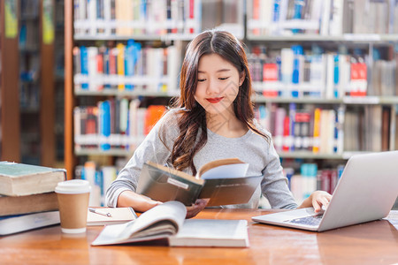 超过做功课和在大学图书馆或使用技术笔记本电脑的亚洲青年学生或具有各种书籍和文具架背景的各种群落返回学校从事家庭作业和使用技术笔记图片