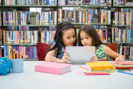 冲浪两个快乐的小可爱女孩在学校的图书馆里玩电脑平板游戏教育和自学无线技术概念人们的生活方式和友爱孩子都喜欢听人说教学生们讲故事应背景图片