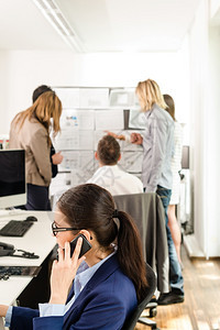 女快乐的一种年轻有吸引力的商业妇女坐在她电脑前用手机坐在她耳边的办公室里在时髦的办公室里与讨论背景概念文件的小组一起讨论背景概念图片