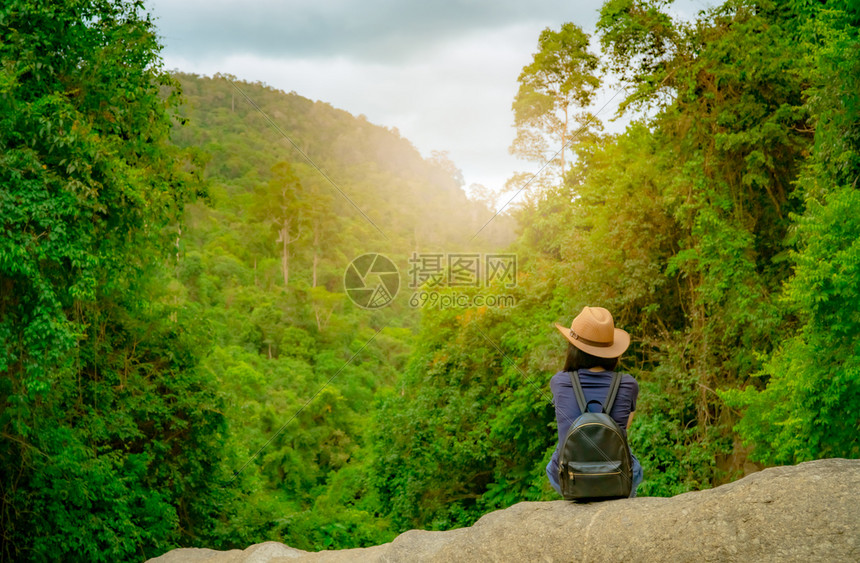 独自的放松夏天快乐女子独自在树林中旅行心情好的活跃女坐在绿谷的石头上森林里有茂密的树快乐假期女子观看着背包聪明的乐队和帽子图片