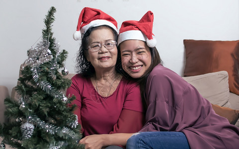 或者高级的两个60岁老母亲和女儿戴着圣塔帽拥抱并坐在家中的客厅里树头模糊圣诞或新年庆典时装瓶子圣诞节或新年庆典概念陈图片