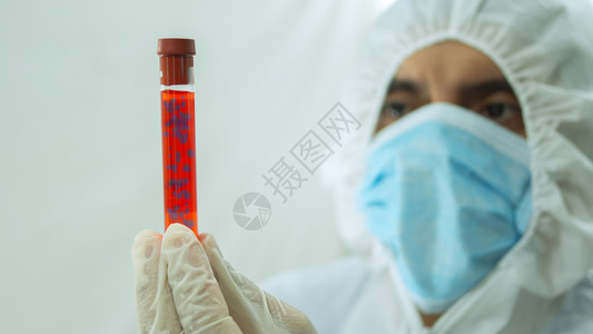 戴着面具和白色生物防护服的医拿着一个带有Covid19标记的试管冠状血液样本用手漂浮在试管内并白色背景上专心地盯着它血样用手漂浮图片