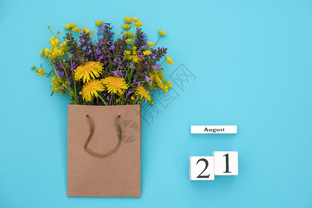 Wooden立方体8月21日历和蓝背景的手工艺品包中富彩的生鲜花蓝色背景的GreetingcardFlatLay概念喜好8月模板图片