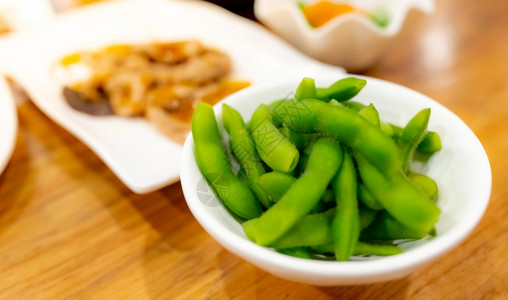 在煮沸前毛豆在日本餐馆木桌上的白碗中浸在饭桌上的Edamame或绿色大豆日美食品模糊的本菜种绿豆高纤维和蛋白质食品基于植物的蛋白质Vegen背景