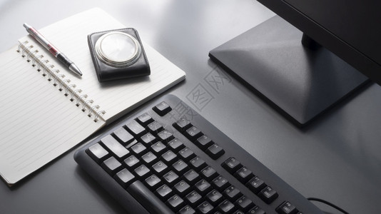 可选择的爱好有地关注黑键盘包括部分计算机监视器圆点笔老式旅行皮革盒手表和灰色桌顶上有阳光和表面阴影的螺旋笔记本白色的图片