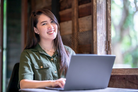 公园坐着学生快乐的亚裔自由职业者商女身着棕褐色绿服装时尚髦的嬉皮节临工使用笔记本电脑与咖啡杯在木屋背景窗口旁工作图片