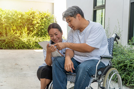 一种支持坐在轮椅上妻子坐在旁边并拿着平板电脑病人因车祸而坐轮椅妻子在旁边让他花园漫步时放松一下不要让他看到与肩并和鼓励的观念健康图片