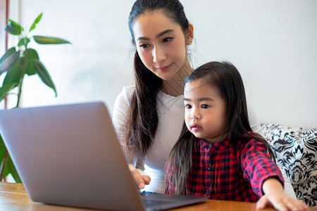 采用浏览幸福亚洲母亲正在教女儿阅读一本书并使用笔记本和技术在家庭学校放假期间进行网上学习教育概念和家庭活动教育概念与家庭活动利用图片