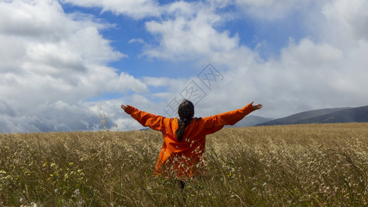 夏天从后面穿橙色夹克的年轻女子身穿橙色外套举起双臂仰望着天空在一片小麦田中央阴云的一天里蓝和山峰在背景中年轻少女从后面穿着橙色夹图片