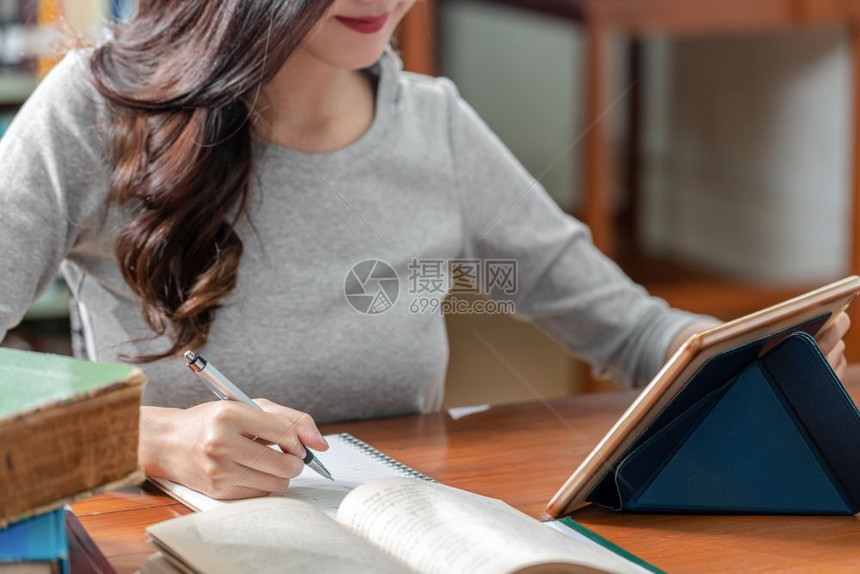 亚洲青年学生手工写功课和在大学图书馆或具有各种本和文具的群落里使用技术平板电脑并记录书架背景返回学校内教育曼谷学习图片