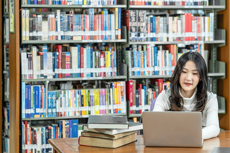 复制日本人做功课和在大学图书馆或使用技术笔记本电脑的亚洲青年学生或具有各种书籍和文具架背景的各种群落返回学校从事家庭作业和使用技图片