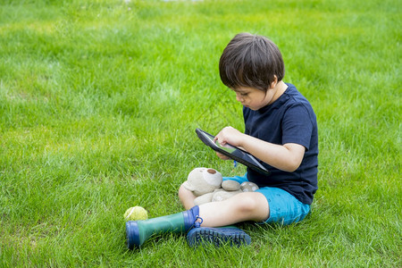 儿童坐在草地上使用平板在春天的花园里做功课在阳光明媚的夏日在家上学社会距离电子学习在线教育中男孩在公园里玩数字垫子孩在线的年轻图片