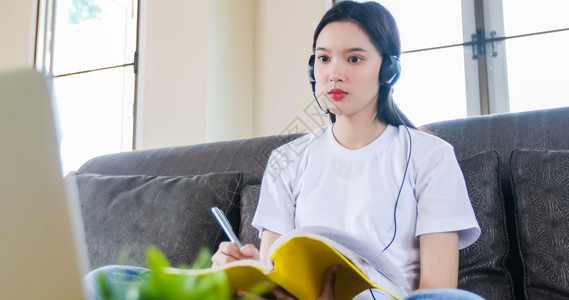 社会的桌子亚裔快乐女学生在网上观看络研讨会播客在笔记式学习教育课程会议上观看网络研讨会播客电话和写笔记看膝上型电脑坐在家中沙发上图片