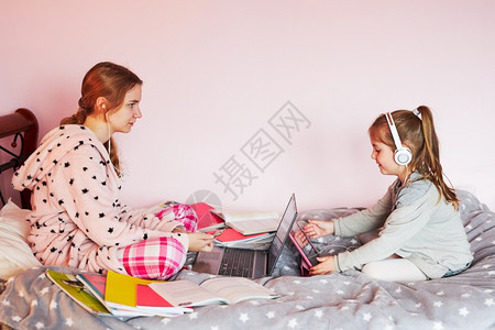 距离手动的学生在线习远程观察课听教授在家中用笔记本电脑耳机书本手册对女孩进行隔离时通过视频电话与在家的同学交谈并坐在床上图片