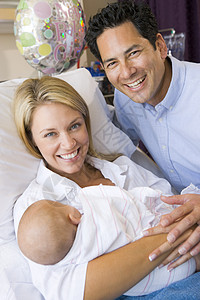新母亲带着婴儿和丈夫在医院中微笑的笑容访问妈妈拥抱花朵气球家长拜访产房三个人相机背景图片