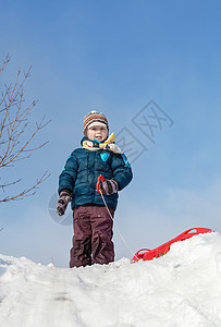在雪山上站着红色塑料雪橇的男孩图片