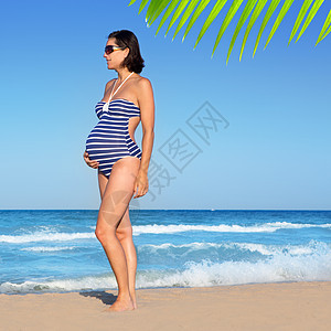 美丽的孕妇站在蓝沙滩上海滩母性父母女性泳装身体成人幸福蓝色支撑图片