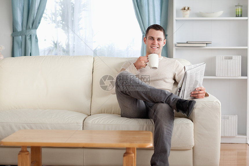 男人在看新闻时喝茶咖啡生活阅读外表闲暇饮料年轻人房间报纸长椅图片
