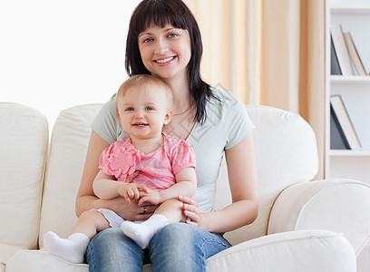 美丽的女人坐在一个女的旁边 抱着她的婴儿在她的怀里休息室喜悦白色儿童生活沙发微笑家庭孩子母亲图片