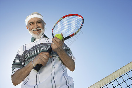 低角度的视角 一个快乐的老年男子 拿着网球花冠和球对着天空图片