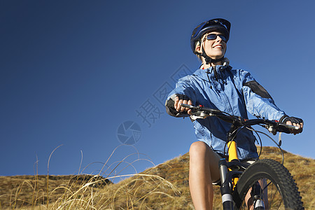 年轻女子坐在田地上骑自行车对抗清蓝天空爱好风衣头盔国家活动成人运动蓝色骑术娱乐图片