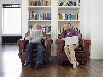 一对中年夫妇在家中用报纸和笔记本电脑坐在手扶椅上坐着的全长中年夫妇快乐硬木地面房间婴儿幸福互联网技术闲暇女士图片