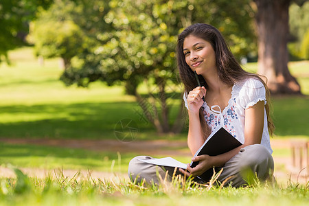 在公园里拿着书和笔的年轻女子长发草地知识分子阅读女士棕色树木花园文学混血图片