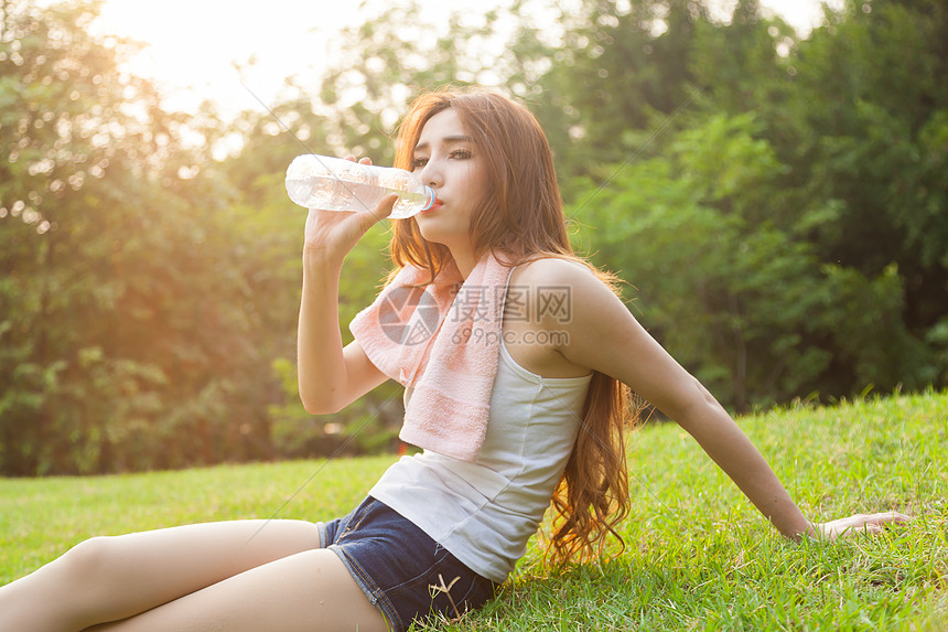 妇女在锻炼后坐着疲劳和饮用水毛巾阳光瓶子运动女士日落草地女孩身体公园图片