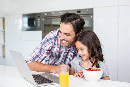 父亲和女儿用笔记本电脑在餐桌上吃早餐时微笑图片
