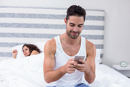 碎屏手机男人在妻子睡觉时使用移动电话 而妻子则睡在背景中背景