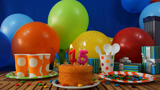 蛋糕蜡烛在土气木桌上的生日蛋糕有五颜六色的气球 礼物 塑料杯子和塑料盘子的背景有糖果和蓝色墙壁的在背景中火焰盒子喜悦幸福桌子庆典蛋糕勺子背景