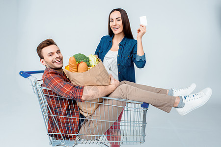 快乐的男子坐在购物车里 拿着杂货袋和笑着女人看空白卡杂货手推车面包购物袋信用卡蔬菜夫妻男人纸袋食物图片