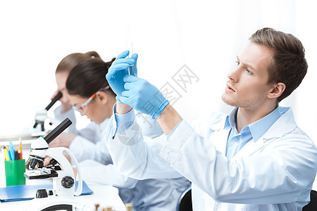 年轻男子化学家在与同事一起坐在桌边时检查试管图片