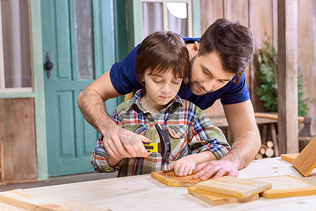 木板上的孩子微笑的父亲教着集中的儿子 在木板上敲钉钉钉背景