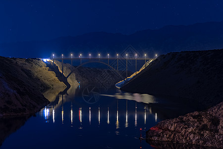 恒星下的闪光高速公路桥 水中的灯光反射 背景的山上和天空中的亮星图片