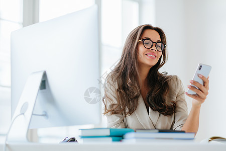 穿着公司服装的微笑女性通过智能手机观看有趣的视频 坐在办公室的桌面上 使用电脑工作 享受工作过程 浏览项目信息 戴眼镜图片