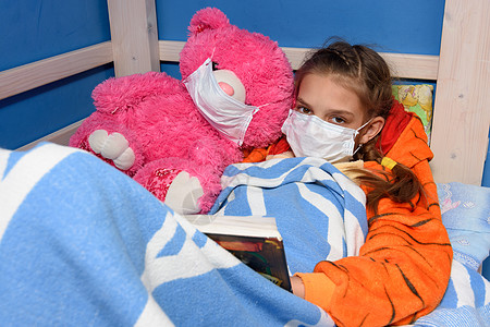 床上玩电脑在床上戴医疗面罩的女童和装在床面药面具中的胖女玩具阅读一本书 并查看框架背景