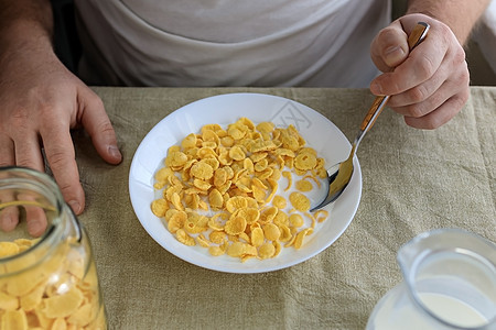 一个剪短的男人拿着一把勺子 勺子放在玉米片里 牛奶放在普通的粗糙桌布上的白盘子里 从上面看 概念 简单快速的美式健康早餐厨房食物图片