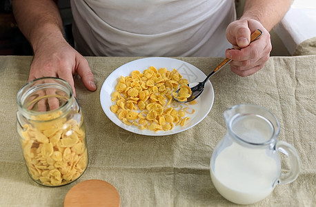 一个剪短的男人拿着一把勺子 勺子放在玉米片里 牛奶放在普通的粗糙桌布上的白盘子里 从上面看 概念 简单快速的美式健康早餐食物男性图片