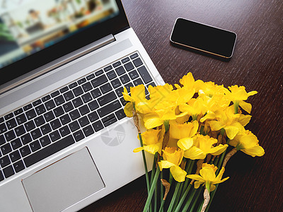 装在银金属笔记本电脑上的Narcissus或水仙 手提装置上的亮黄色花朵 木制背景的智能手机职场工作花束水仙花花瓣工具桌子植物商背景图片