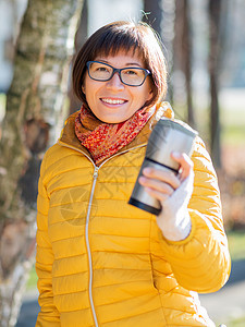 穿着亮黄色夹克的快乐笑容的女人拿着热水杯 在寒冷的秋天喝热茶或其他饮料咖啡旅游女孩眼镜女士烧瓶游客公园女性旅行图片