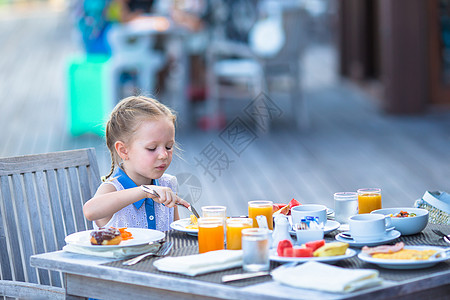 可爱的小女孩在户外咖啡厅吃早餐眼睛蜂蜜桌子食物金发瓶子橙子水果婴儿女孩图片